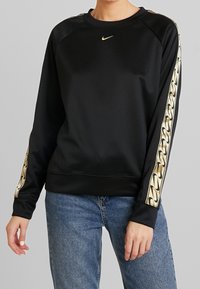 Svart Nike-tröja med en slät textur, rundad halsringning och guldkedjemönstrade detaljer på ärmarna. Kommer tillsammans med blå jeans.