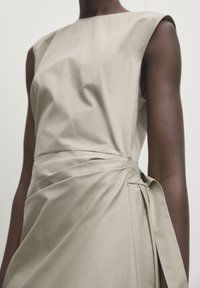 Robe sans manches beige avec un détail de taille froncée fermée par un bouton noir sur un modèle à la peau noire, sur fond uni.