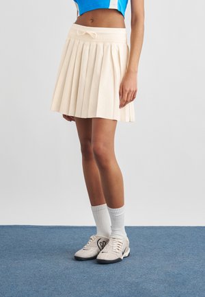 Mujer con falda plisada color crema, calcetines blancos, zapatillas beige y top corto azul, de pie sobre una alfombra azul contra un fondo liso.