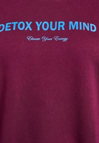 Bordová T-shirt s modrým textom "DETOX YOUR MIND" je výrazne zobrazený. Nižšie, v menšom písmene, je napísané "Elevate Your Energy." Mäkka textúra.
