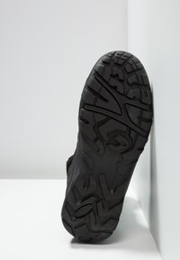 Black sneaker sole with deep treads, featuring a textured surface and abstract design on the outer sole, resting against a wall.
