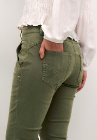 Jeans de culoare verde măsliniu, cu un design ajustat și detalii la buzunarul din spate. Materialul pare neted, cu accente subtile de cusătură și butoane metalice mici.