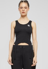 Svart tettsittende tanktopp med utskjæringsdesign i midjen, laget av myk stoff, med en liten logo på brystet og brede stropper.