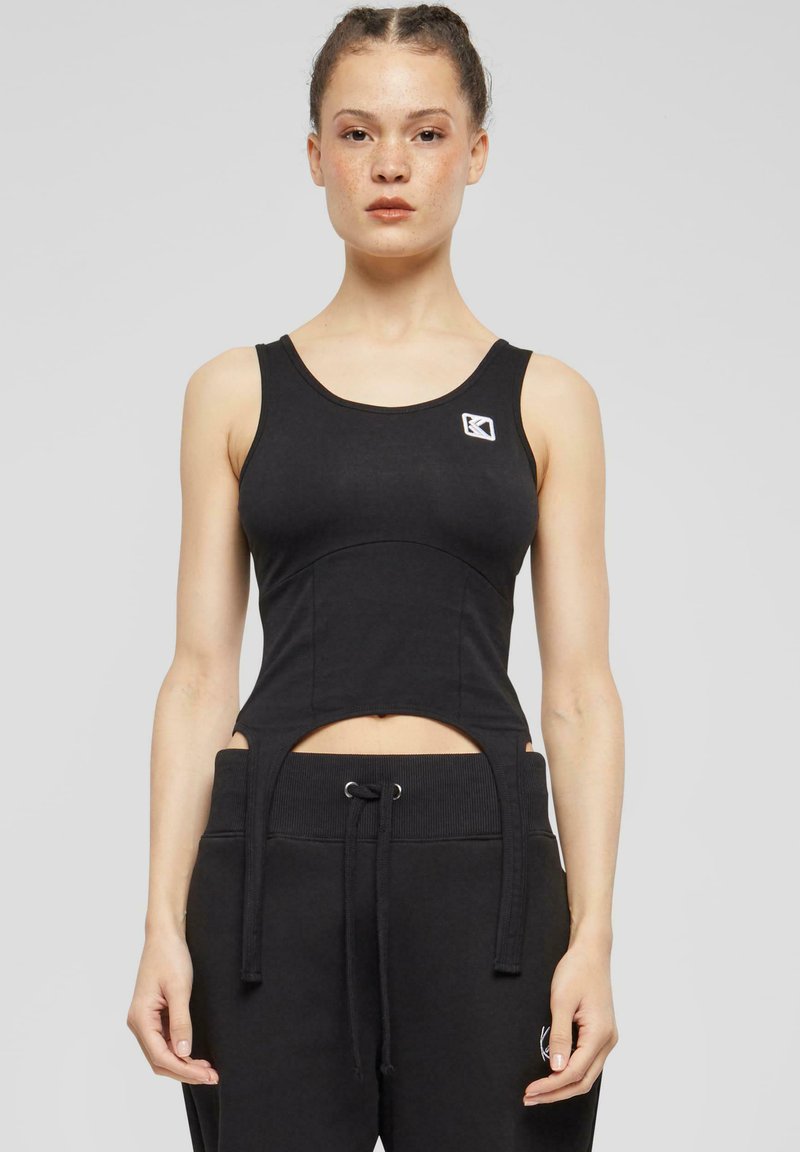 Svart tettsittende tanktopp med utskjæringsdesign i midjen, laget av myk stoff, med en liten logo på brystet og brede stropper.
