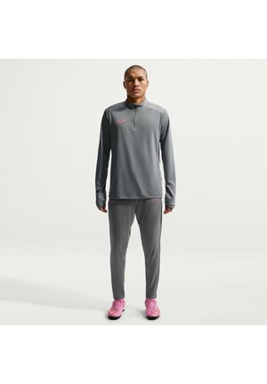 Osoba stoi wyprostowana, ubrana w pasujący szary dres Nike z różowymi logotypami i różowymi butami sportowymi na prostym jasnym tle.