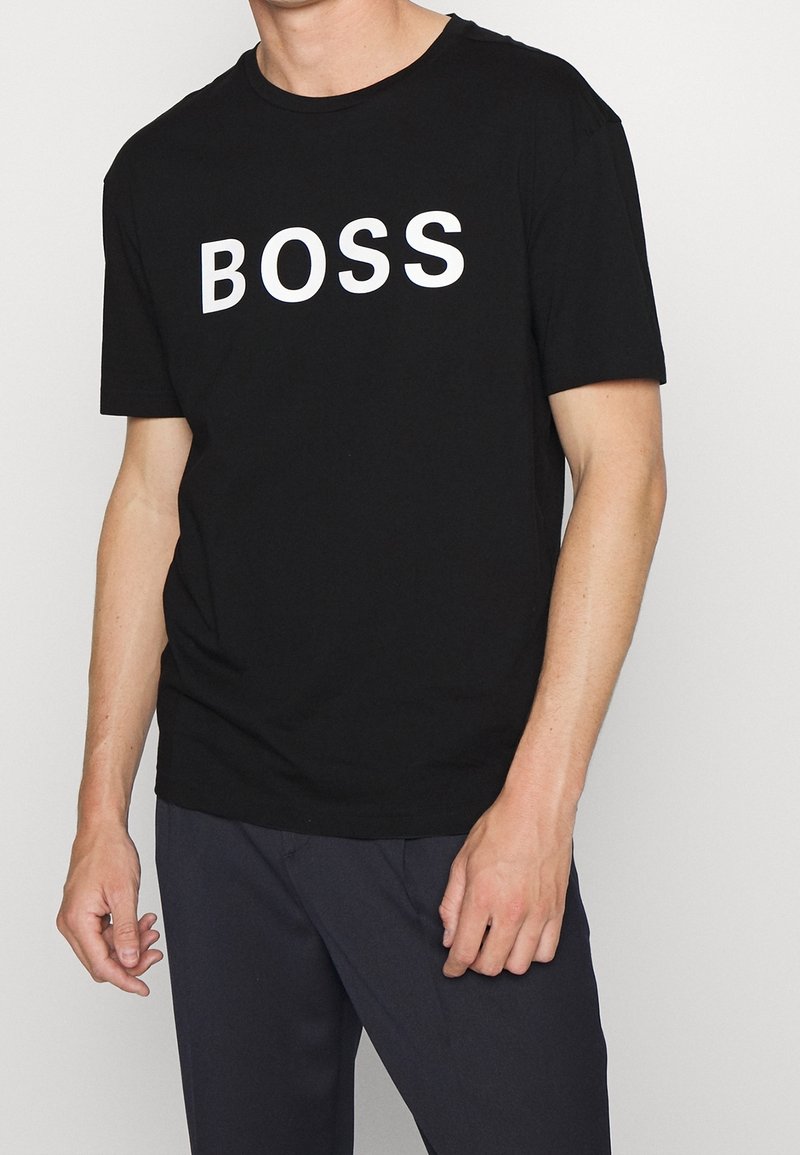 Homme portant une chemise noire à manches courtes avec le mot « BOSS » en grandes lettres blanches sur la poitrine, associé à un pantalon foncé.