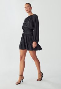 Crna krepa haljina s dugim rukavima, nabranu bodice, strukiranim strukom i slojevitom suknjom. Uparena s crnim sandalama s remenčićima.