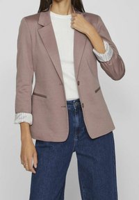 Veste rose à coupe simple boutonnage, tissu texturé, avec poignets contrastants à motifs. Associée à un jean bleu foncé.