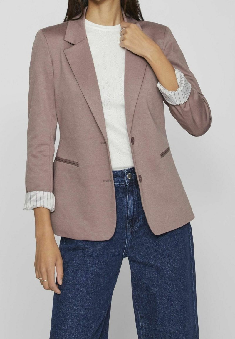 Veste rose à coupe simple boutonnage, tissu texturé, avec poignets contrastants à motifs. Associée à un jean bleu foncé.