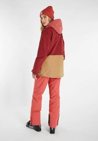 Rode en tan waterdichte jas met een capuchon, gecombineerd met roze sneeuwbroek. Bevat ritsen bij de mouwen, wat een sportieve winterstijl weerspiegelt.