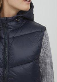 Die marineblaue Steppweste hat einen hohen Kragen und einen Reißverschluss. Die Außenseite ist glatt und glänzend, mit einem grauen Strickpullover, der leicht sichtbar darunter getragen wird.