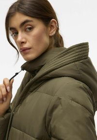 Ženska prilagaja zadrgo olivno zelene jakne s prešito teksturo in velikim podloženim kapuco, nosi več srebrnih uhakov.