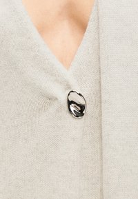 Κοντινό πλάνο μπεζ πλεκτής ζακέτας με βαθύ λαιμόκοψη σε σχήμα V και μεγάλο λαμπερό ασημένιο κουμπί που κλείνει το μπροστινό μέρος.