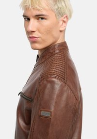 Braune Lederjacke mit strukturierten Schulterakzenten, einer Reißverschlusstasche auf der Brust und einer glatten Oberfläche; verfügt über einen Stehkragen und dezente Markenkennzeichnung.