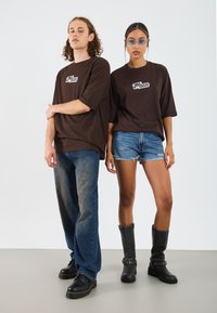 Camisetas oversized marrones con logo, pantalones cortos de mezclilla azules, botas negras por el tobillo y botas altas negras. Texturas suaves y estilo casual.