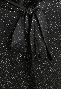 Tejido negro con lunares blancos, que presenta un detalle de lazo atado en la cintura, hecho de un material suave y liso.