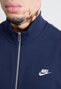 Casaco azul marinho com fecho éclair, feito de algodão e com colarinho alto. Apresenta um fecho éclair prateado e um logótipo branco da Nike na área do peito esquerdo.