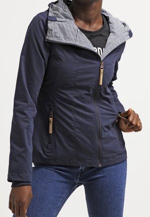 Person iført en navyblå jakke med lynlås, stribet indvendig hætte og lynlåslommer, kombineret med blå jeans og en sort skjorte under.