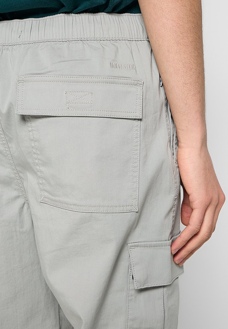 Helle graue Cargo-Hose mit elastischem Bund, zwei aufgenähten Taschen auf der Rückseite und einer Seitentasche mit Reißverschlusdetail. Glatte Stofftextur.