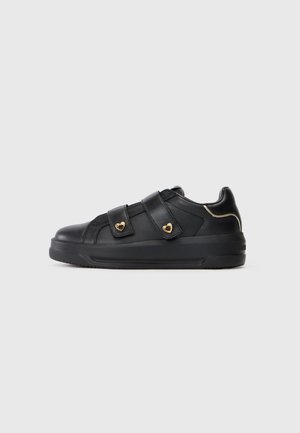 Zapatillas negras de cuero vegano con dos correas de Velcro y acentos en forma de corazón dorados. Presenta una suela de goma gruesa y un diseño minimalista.