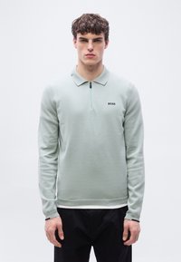 Νεαρός με σγουρά μαλλιά, φορώντας ένα ανοιχτοπράσινο πουλόβερ με φερμουάρ και το λογότυπο "BOSS" και μαύρο παντελόνι, στέκεται μπροστά σε ένα απλό λευκό φόντο.