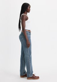 Lichtblauwe denim jeans met een rechte pasvorm, voorzien van een vijf-pocket ontwerp en gestikte achterzakken, gecombineerd met bruine schoenen en een witte tanktop.