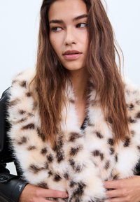 Faux fur krave i crème med mørkebrune leopardpletter. Blød tekstur og voluminøst design med en bred, fyldig kant. Sort læderjakke synlig.