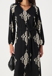 Vestido negro de manga larga, con escote en V, que presenta un patrón ornamentado en crema. Incluye un cinturón a juego y está hecho de una tela suave y fluida.