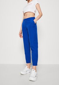Femme portant un pantalon bleu vif fuselé, un crop top blanc et des baskets Nike blanches, debout devant un fond blanc uni.