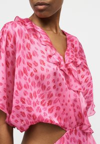 Γυναίκα που φοράει ροζ σατέν μπλούζα με κόκκινο εκτύπωμα χειλιών και βολάν στο λαιμό σε σχήμα V, κομμένη ώστε να φαίνεται ο άνω κορμός και το ντεκολτέ.