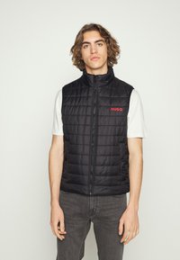 Gilet trapuntato nero con colletto alto e zip frontale, con il logo rosso "HUGO". Indossato sopra una maglietta bianca, abbinato a jeans scuri.