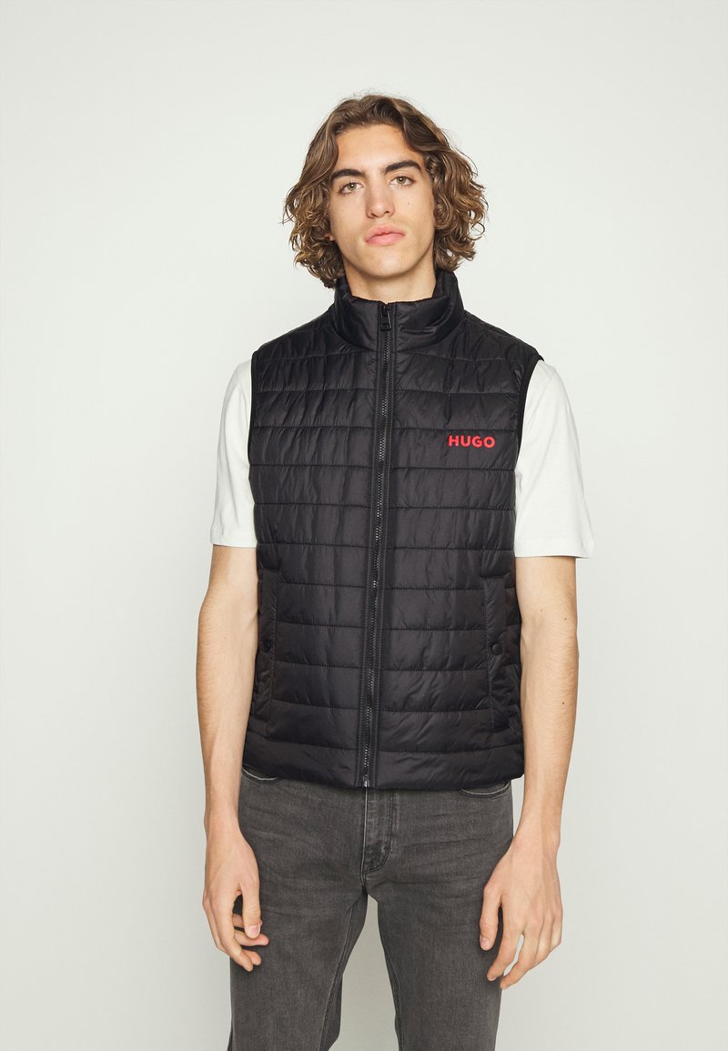 Gilet trapuntato nero con colletto alto e zip frontale, con il logo rosso "HUGO". Indossato sopra una maglietta bianca, abbinato a jeans scuri.