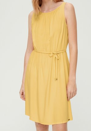 Femme portant une robe jaune sans manches, longueur genou, avec une fine ceinture nouée à la taille, sur un fond clair uni.