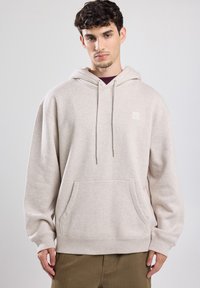 Beige Kapuzenpullover aus weichem Stoff, mit einer vorderen Kängurutasche, einer verstellbaren Kordelzugkapuze und kleinem Logo auf der Brust.