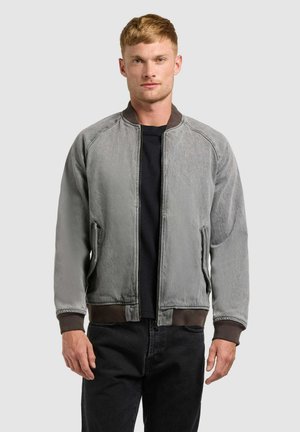 Fiatal férfi, aki kopott szürke bomber dzsekit visel bordázott mandzsettával és derékpánttal, egyszínű világos háttér előtt áll.