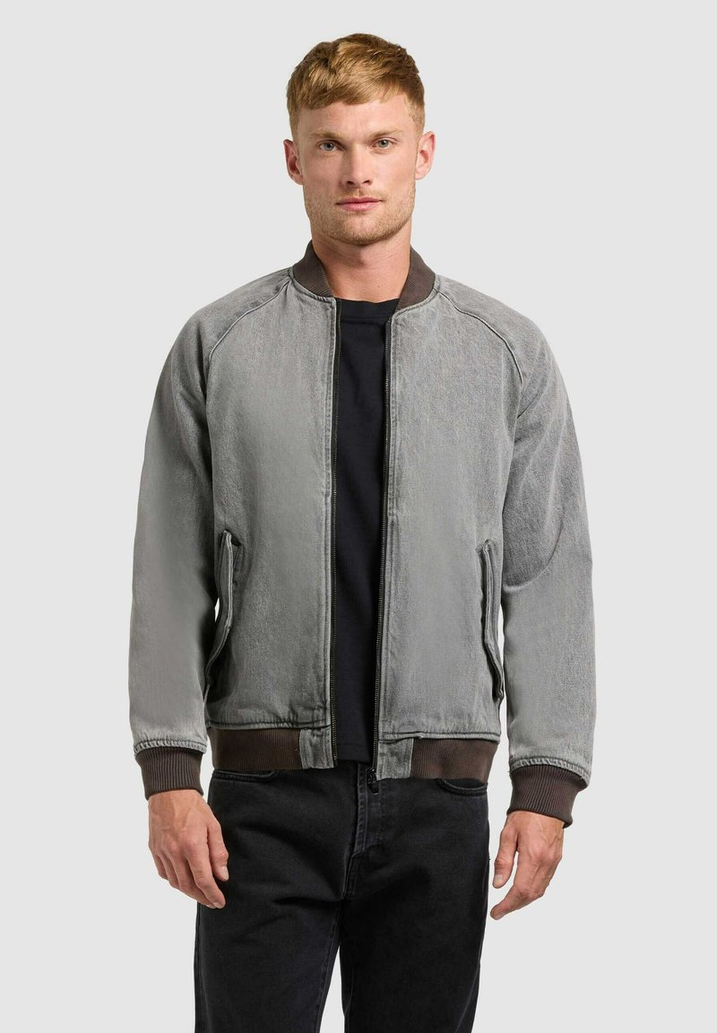 Jeune homme portant une veste bomber grise délavée avec des poignets et une taille côtelés, debout devant un fond clair uni.