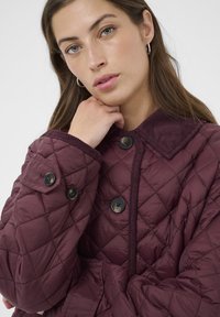 Bordeauxfarbene gesteppte Jacke mit Druckknopfverschlüssen, einem Cordkragen und strukturiertem Stoff mit Rautenmuster. Weiches und warmes Material.