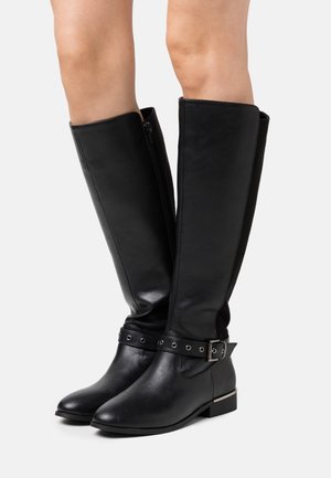 Bottes hautes en cuir noir avec fermeture éclair latérale et sangle décorative avec boucle, portées sur jambes nues sur fond blanc.