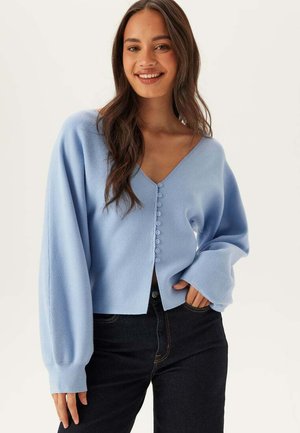 Ung kvinde smilende, iført lyseblå knap-op cardigan med lange ærmer og sorte bukser, stående foran en ensfarvet hvid baggrund.