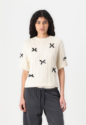Maglione di cotone crudo a maglia cable con maniche corte, decorato con fiocchi neri in raso, con colletto e orlo a coste. Jogger grigi visibili sotto.