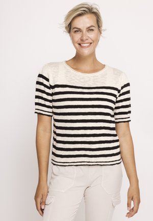 Smiling woman with short blonde hair wearing a black and white striped short-sleeve shirt and light-colored pants against a plain background.