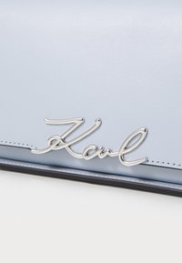 Světle modrý kožený povrch s metalickým stříbrným křivým logem "Karl" připevněným blízko prošitého okraje.