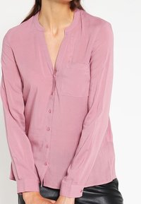 Blouse rose boutonnée avec col mandarin, manches longues, poche avant, et tissu doux et texturé.
