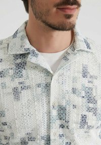 Chemise à manches courtes en bleu clair et blanc, arborant un motif texturé avec des formes géométriques et de petits boutons le long de la patte de boutonnage.