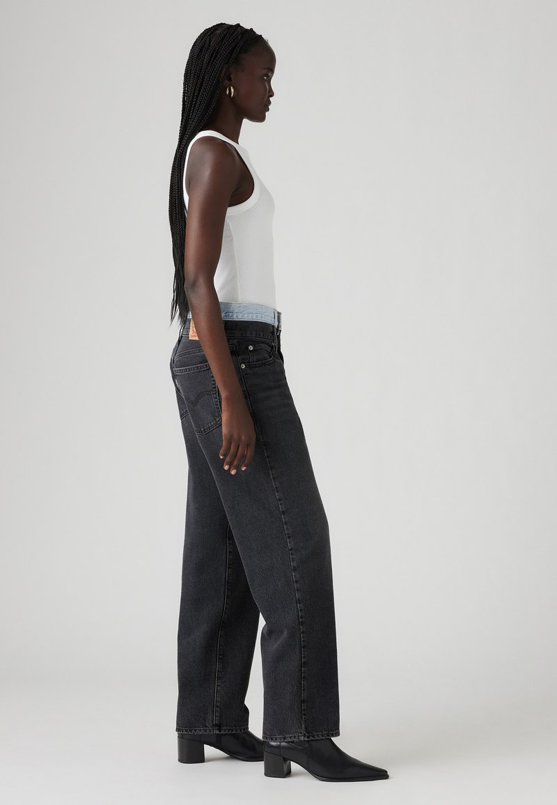 Jean noir taille haute à coupe droite, avec un détail de ceinture bleu clair, associé à un débardeur blanc et des bottines à talons noires.