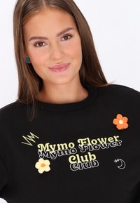 Fekete pulóver zöld és fehér hímzéssel, melyen a "Mymo Flower Club" felirat olvasható, sárga és narancssárga virág-applikációkkal díszítve.