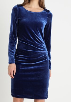 Femme portant une robe en velours moulante à manches longues, longueur genou, de couleur bleu profond, avec un col rond et un détail froncé à la taille.