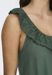 Zelena bezrukavna majica s teksturiranim volanom s rupicama, izrađena od lagane tkanine s glatkom završnom obradom.