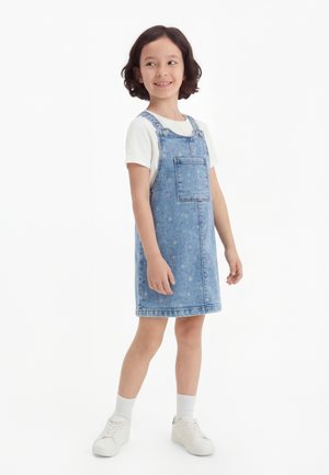 Młoda dziewczynka w białej koszulce, dżinsowej sukience z małymi kolorowymi nadrukami, białych skarpetkach i białych trampkach, stoi i się uśmiecha.