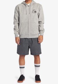 Sudadera gris con cremallera y logo, combinada con pantalones cortos oscuros. El modelo lleva zapatos negros y calcetines blancos con rayas negras y azules. Atuendo casual.